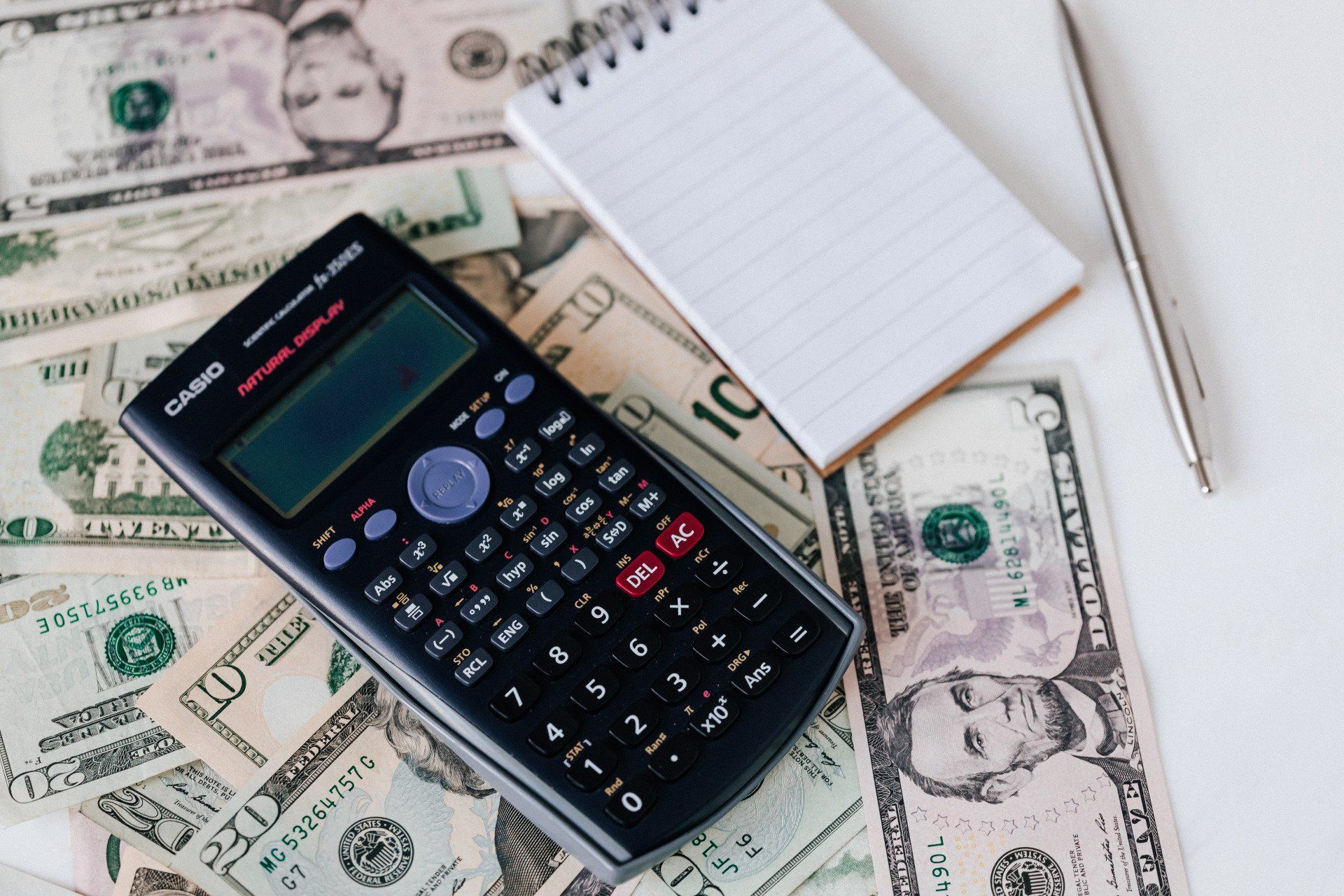 Calculator on U.S. dollar bills beside a notepad and pen on a white surface
